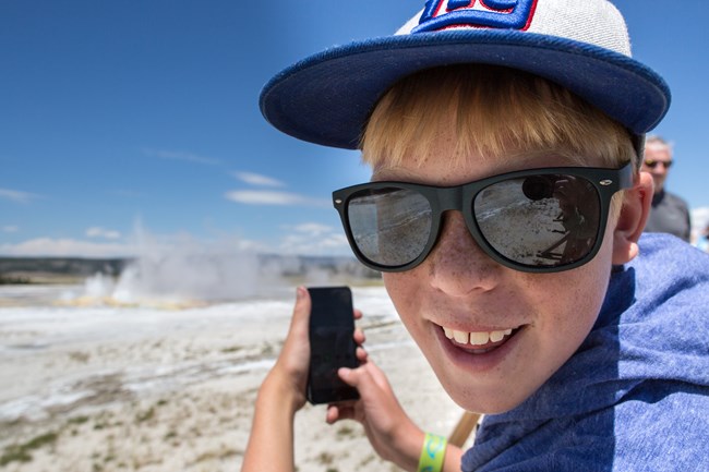 a boy in sunglasses and a hat looks over his shoulder and smiles while taking a photo of the scenery