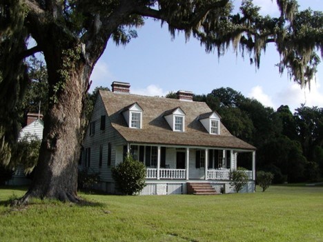 A white house stands next to a large tree on a green lawn. Green trees line the back of the house.