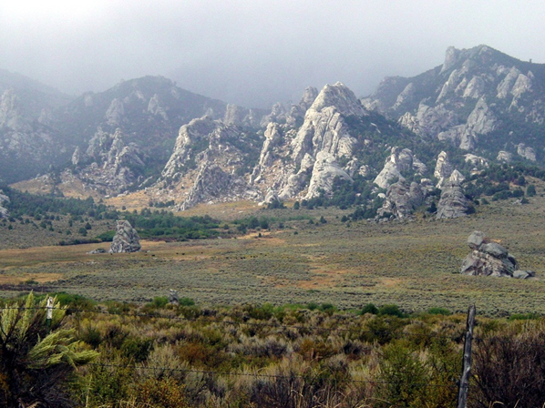 Sagebrush plains lie in the foreground.  A wall of granite domes and spires line the background. The sky is foggy with snow.