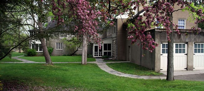 A large brown house with many white doors and windows, walkways, and trees, including some with bright pink flowers.