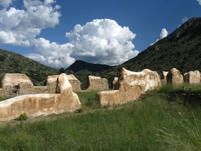 Weathered, tan-colored rock structures sit amongst green grasses, in front of low mountains. The blue sky has white, puffy clouds. Weathered, tan-colored rock structures sit amongst green grasses, in front of low mountains. The blue sky has white, puffy clouds.