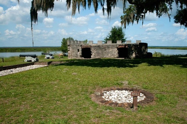 The remains of a stone building are flanked by artificial cannons on the left. The building is surrounded by water in the background; the remains of a water well are in the foreground.