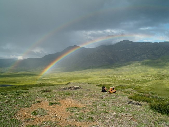 A double rainbow arches over a green river valley. Mountains are in the background and the sky is darkened by clouds.