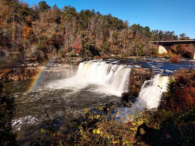 A large waterfall creates a rainbow as it drops into the river below. Autumn foliage, with many leaves already having fallen, surrounds the waterfall.