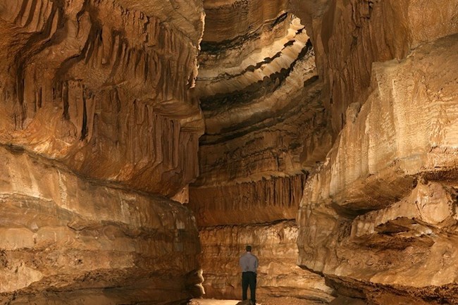 A park ranger is dwarfed by the reddish rocks of a large cave. A park ranger is dwarfed by the reddish rocks of a large cave.
