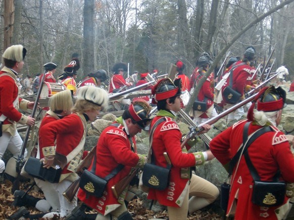 A group of men dressed in red militia uniforms kneel behind a stone wall during battle. Smoke fills the air, filtering the wooded background.