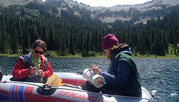MORA-2015-350-x-200 Image of two individuals handling scientific instruments in a raft floating on a lake.