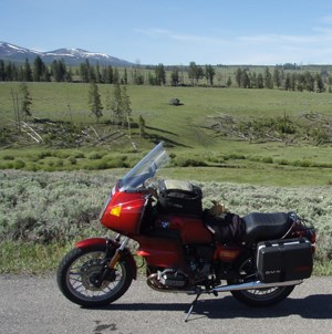 lt= Red motorcycle parked on roadside with meadow, trees, and mountains in background.