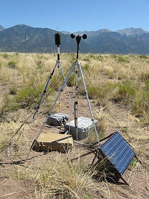 Microphone, weather station, and solar panel in dry landscape with mountains in the background.