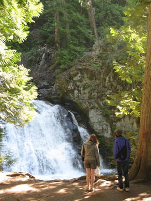 lt= Two people stand in front of a large waterfall in the woods.
