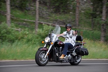 lt= Man wearing helmet riding a motorcycle on a road through the woods.