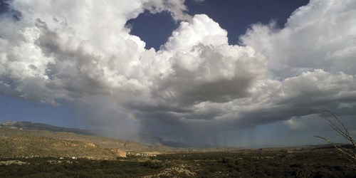 Large storm clouds rain down on a desert landscape, with a rainbow visible