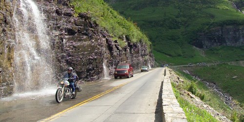 Motorcycle driving up a steep road, with several vehicles behind and a waterfall to the left