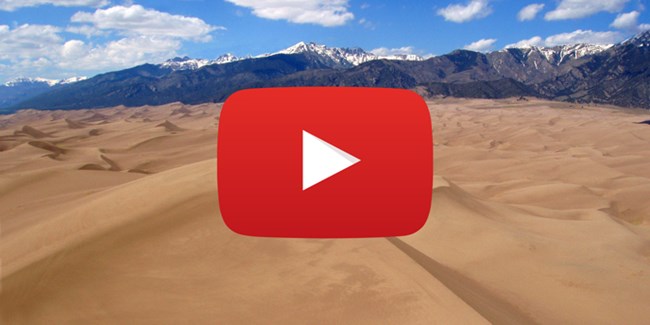 YouTube play logo over a field of sand dunes with mountains and blue sky in the background. YouTube play logo over a field of sand dunes with mountains and blue sky in the background.