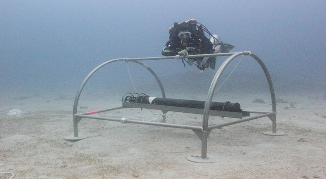 Underwater ocean view of a scuba diver inspecting a hydrophone on the ocean floor.