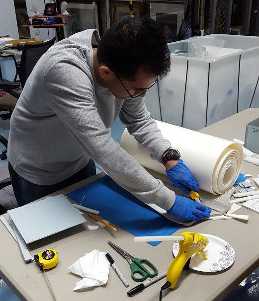 Man with brown skin and hair wearing glasses and gloves works on a project with paper tools and scissors