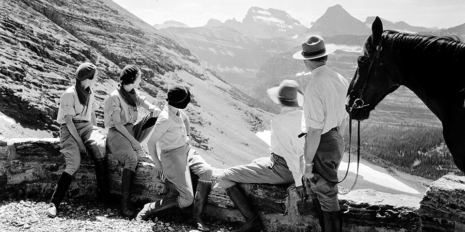 historic black and white photo of three women wearing period clothing sitting on a rock wall next to a standing park ranger and horse looking at distant mountains.