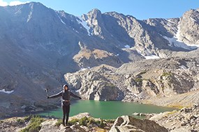 A hiker stands with outstretched arms, behind her a blue lake in the distance
