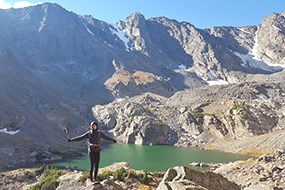 A hiker stands with outstretched arms, behind her a blue lake in the distance A hiker stands with outstretched arms, behind her a blue lake in the distance