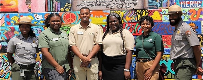 Group of six African American people standing in front of a wall, two are park rangers on either side and between them are college students