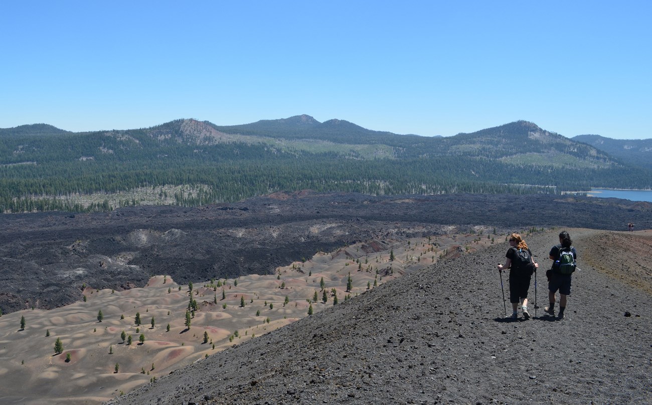 Photo of 2 people walking on cinder cone trail overlooking painted dunes, fantastic lava flow, butte lake, and several low forested peaks.