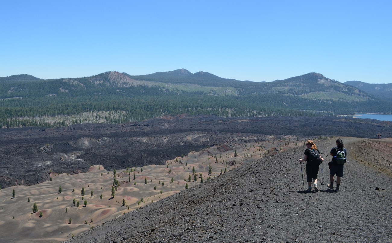 9dbf42d3-8cd9-49f0-b7ac-3261ff04c618Original-LAVO-webcrop Photo of 2 people walking on cinder cone trail overlooking painted dunes, fantastic lava flow, butte lake, and several low forested peaks.