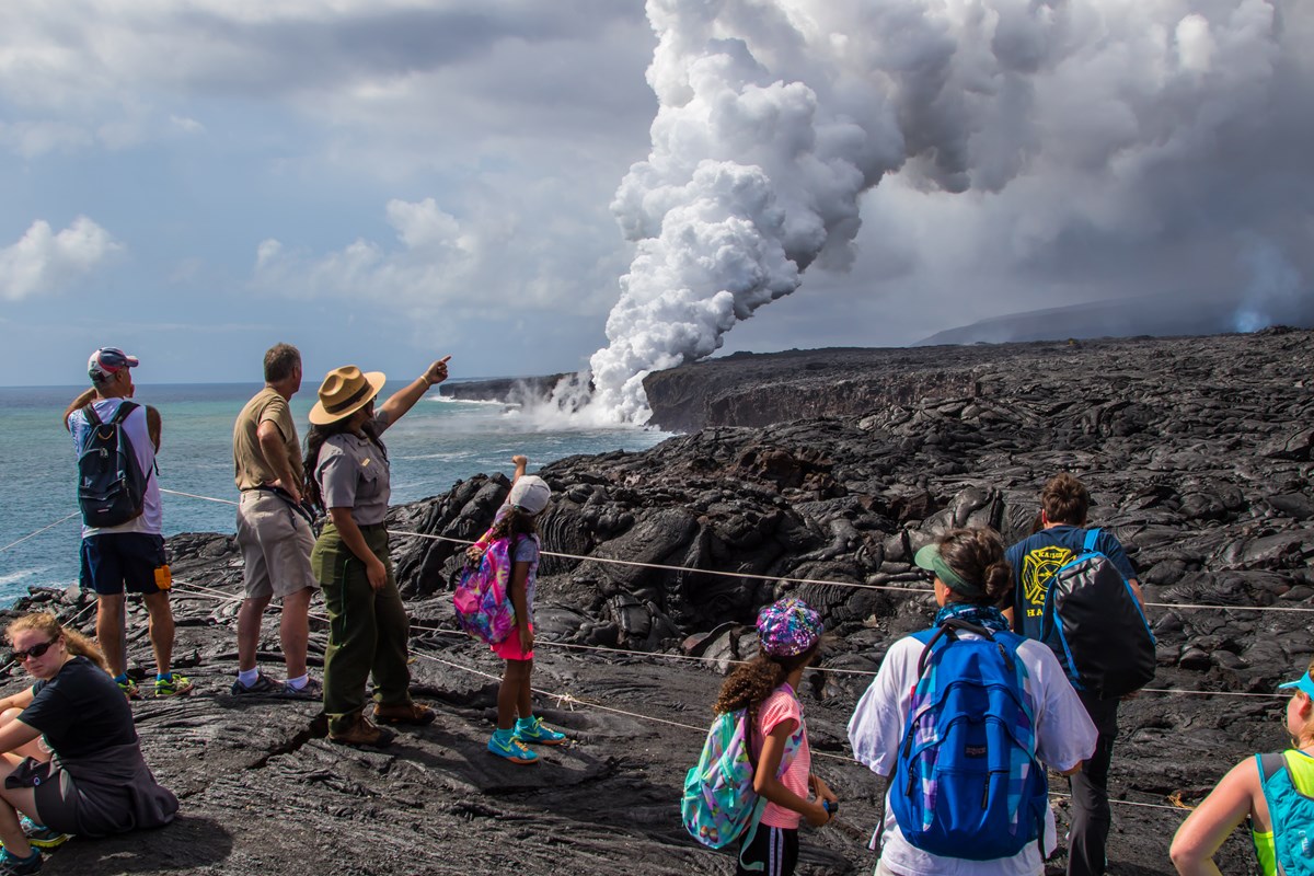 park ranger talking with people at overlook of lava field with steam plume where molten lava enters the ocean