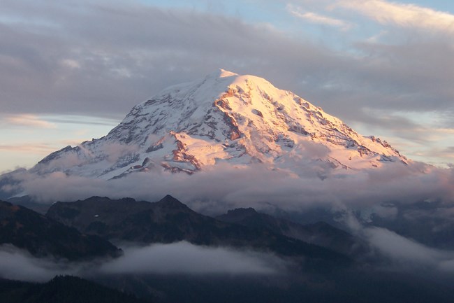 Mount Rainier towers over all surrounding mountains, lit by the sunset and wreathed in clouds.