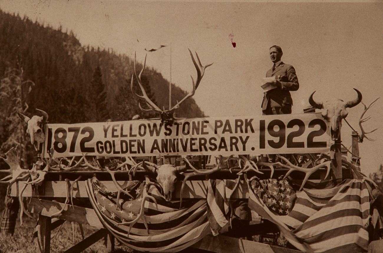 YELL centennial NPS Director Horace Alright givigin a speech from a stage with a sign celebrating the Yellowstone Golden Anniversary in 1922.
