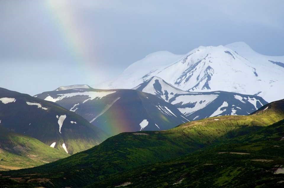 photo of hills with tundra vegetation and a snow covered peak in the distance behind a colorful rainbow