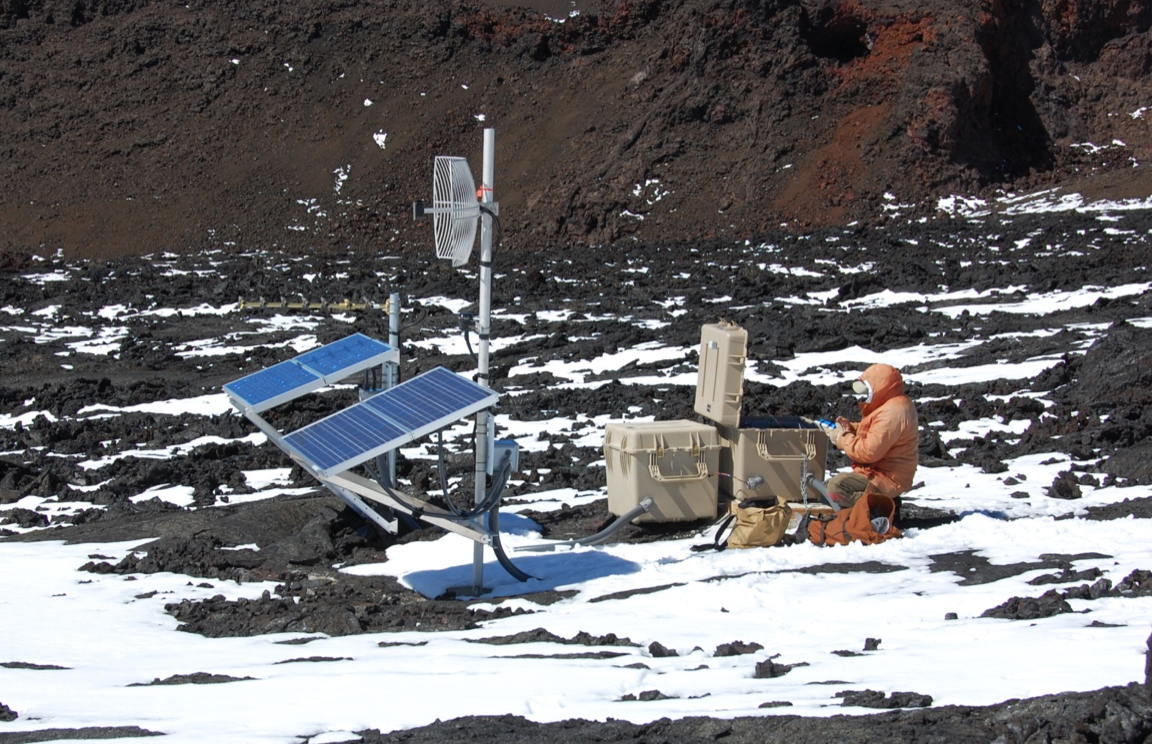 seismic station vhp_img5109 Photo of a person sitting with monitoring instruments on a snowy mountainside.