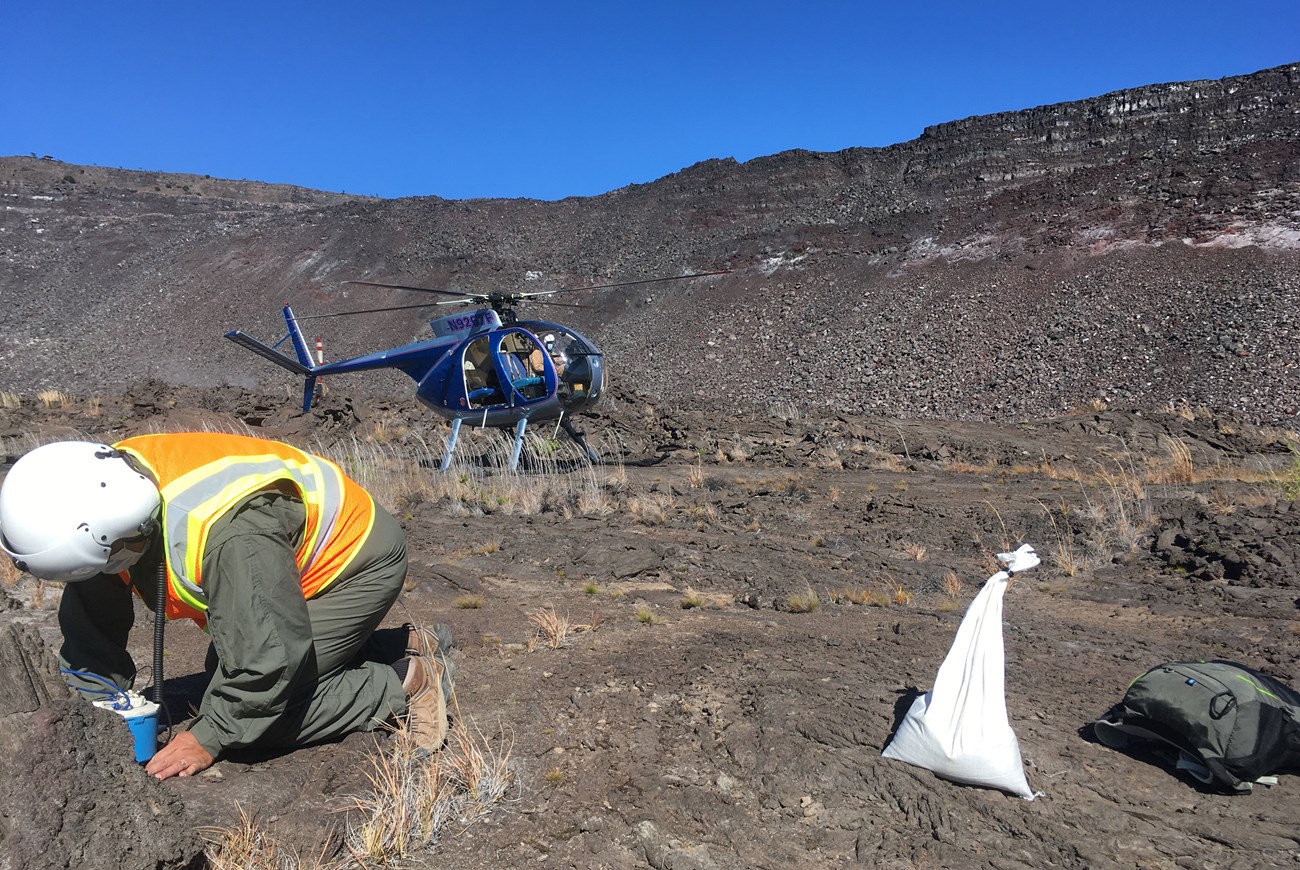 usgs-A3_Nodel_Deployment_DDB-web photo of a person wearing a flight suit and helmet working in a lava field with a helicopter on the ground in the distance