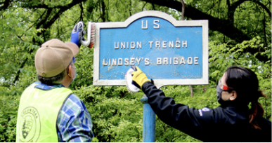 A photo of Kristyn, a Community Volunteer Ambassador from Vicksburg National Military Park, cleaning a sign with a volunteer that reads “U.S. Union Trench Lindey’s Brigade”