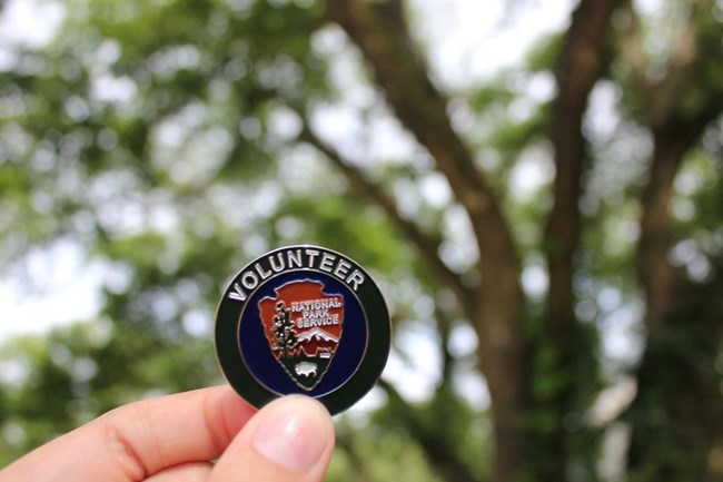 A close-up photo of a dollar-sized metal coin of the National Park Service Volunteers-In-Parks program logo, including white text that reads "Volunteer" above an inner circle with the NPS arrowhead.