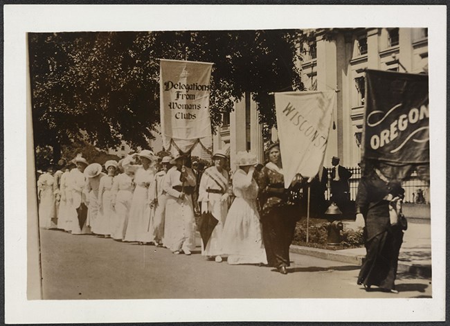 Photograph of contingents of suffragists marching with banners on street. Banners read: "Delegations From Womans Clubs", "Wisconsin," and "Oregon."