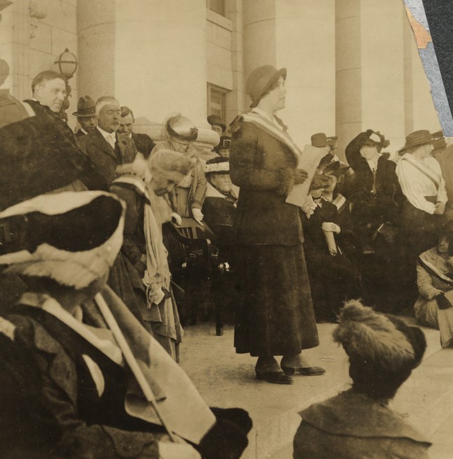 1915 photo of a woman wearing a suffrage sash speaking in front of a group of people