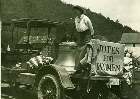 black and white photo of woman standing on the flatbed of an early 20th century car next to a large bell and a sign "Votes for Women'
