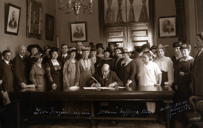 a black and white image of a man at a large desk signing a document surrounded by a group of women