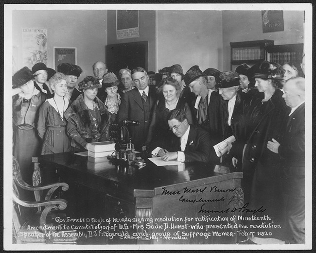 black and white photo of a man at a desk signing a document surrounded by women