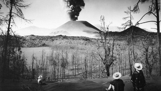 Black and white photo of three people in broad hats watching a distant volcanic eruption