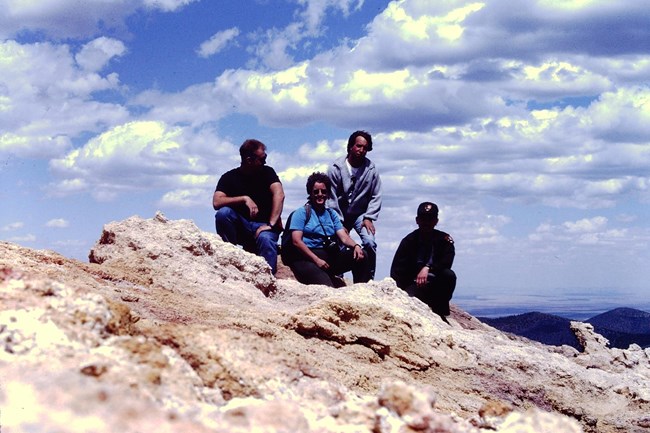 Four people sit on yellow-white rocks high above surrounding desert and forest terrain. One of them is a park ranger. They group is on top of Sunset Crater Volcano. White clouds dot the blue sky above them.