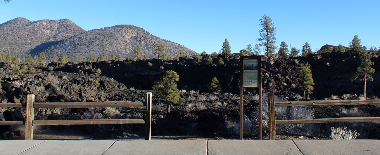 A trailhead sign stands by a wooden fence along a sidewalk. Black jagged rocks, scattered trees, and a mountain are in the background