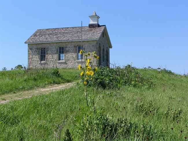 stone building at top of hill and road surrounded by green prairie, blue sky