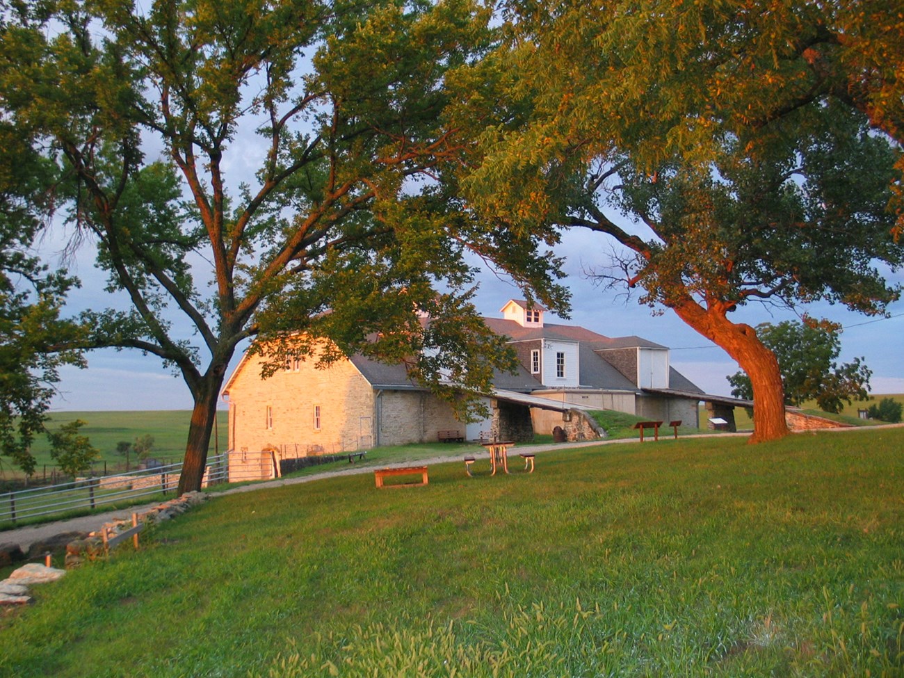 Lawn with picnic table, trees and road with limestone barn behind at sunrise
