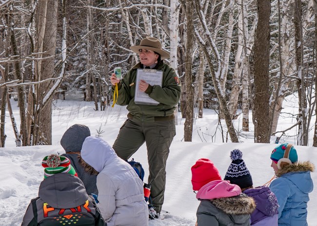 A ranger holds a small laminated piece of paper surrounded by chikdren sitting in the snow in the woods
