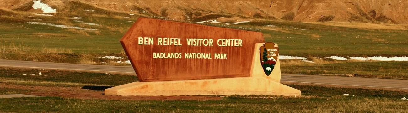 a sign reading ben reifel visitor center sits in front of a road and badlands formations, all bathed in a warm sunset glow.