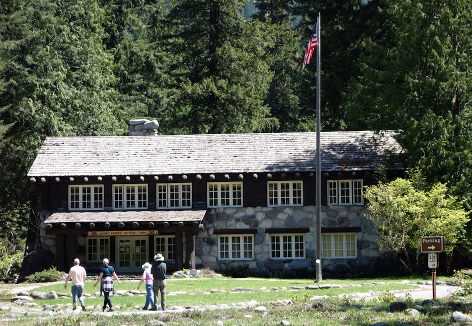 Several people walk along a path towards a two story wood and rock building surrounded by forest. Several people walk along a path towards a two story wood and rock building surrounded by forest.