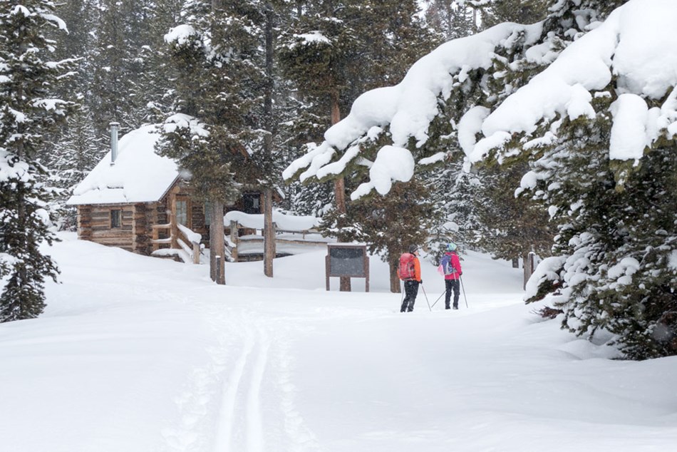 Two skiers wait near the log registration building along the Indian Creek Loop Ski Trail. Two skiers wait near the log registration building along the Indian Creek Loop Ski Trail.