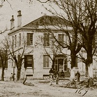 two-story house with closed shutters surrounded by trees