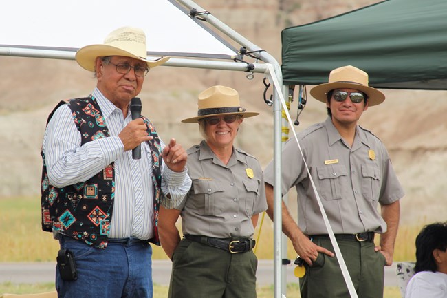 a lakota man wearing a beaded vest speaks into a microphone with two park rangers looking on and smiling.